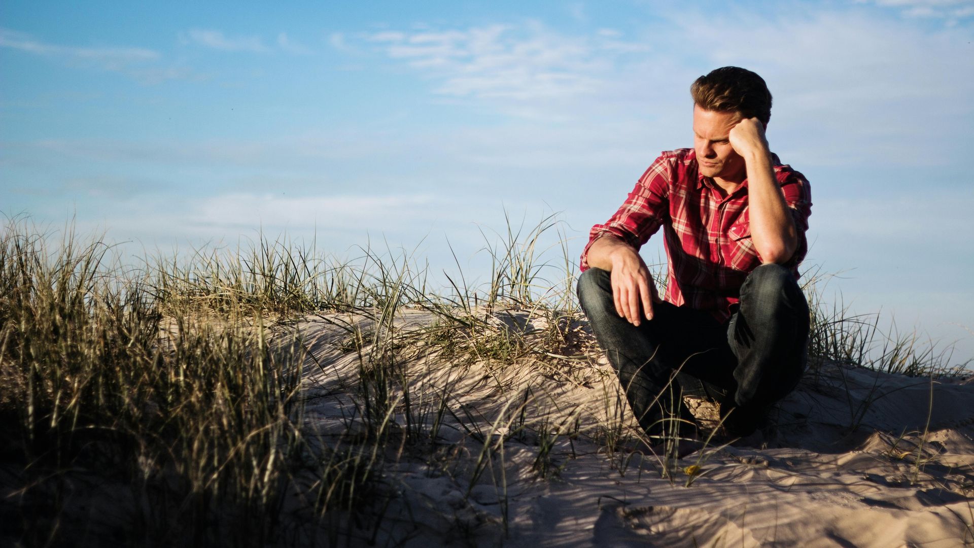 Person sitting on sand dunes, wearing a red plaid shirt, looking down under a clear blue sky