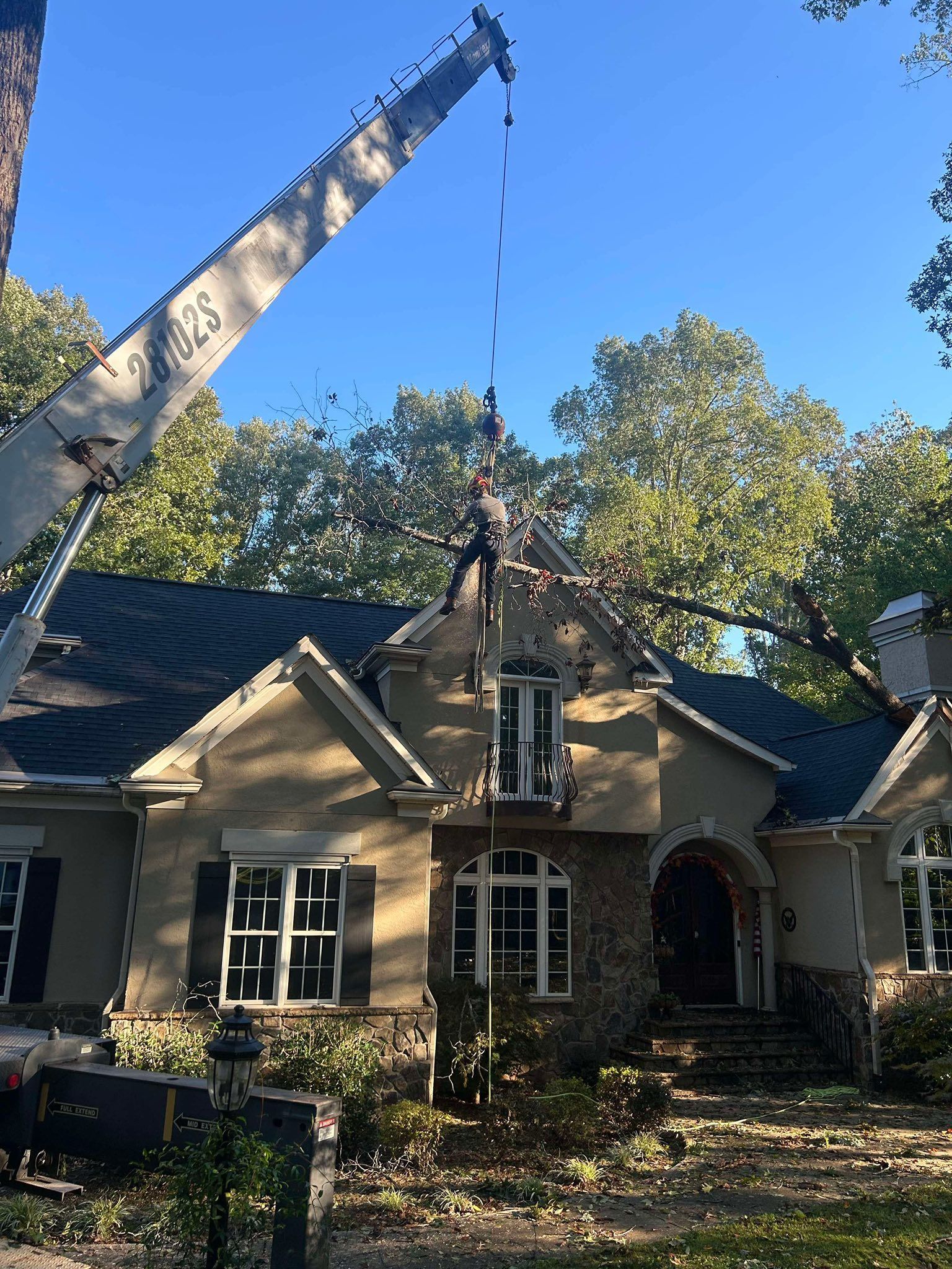 A crane lifts a large tree limb away from the roof of a beige house with a dark shingled roof and stone accents.
