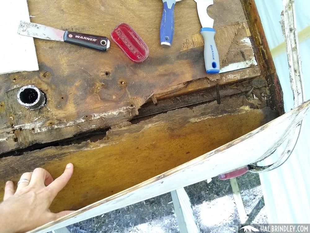 A person's finger points to damaged, water-stained wood in a trailer, tools are nearby, indicating repair.