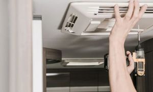 A person is using a power drill to install or repair an RV air conditioner. Interior setting with neutral tones.