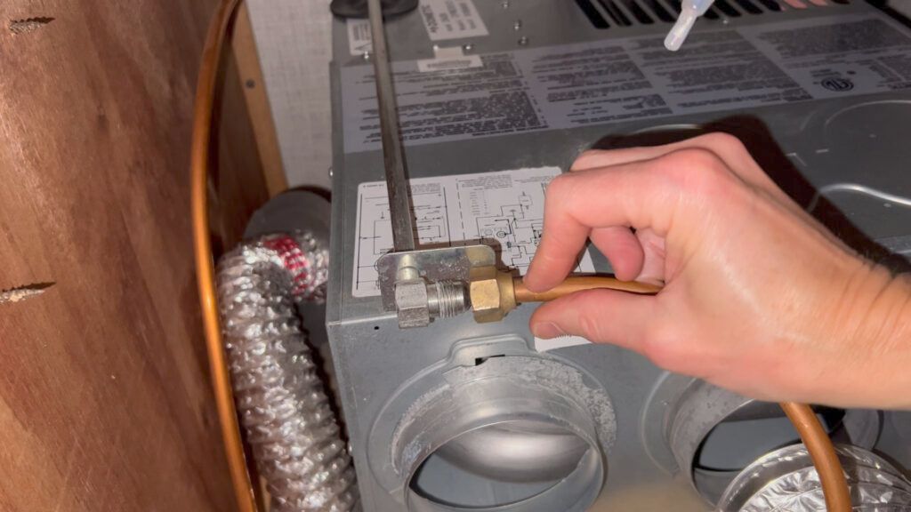 A person's hand connecting a copper pipe to a brass fitting on a metal furnace.  The setting is indoors.