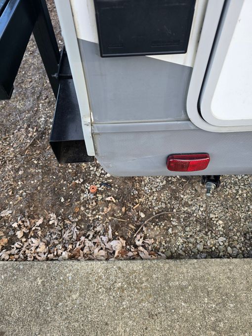Lower corner of a white and gray RV showing a red taillight, black metal bracket, and gray trim. 