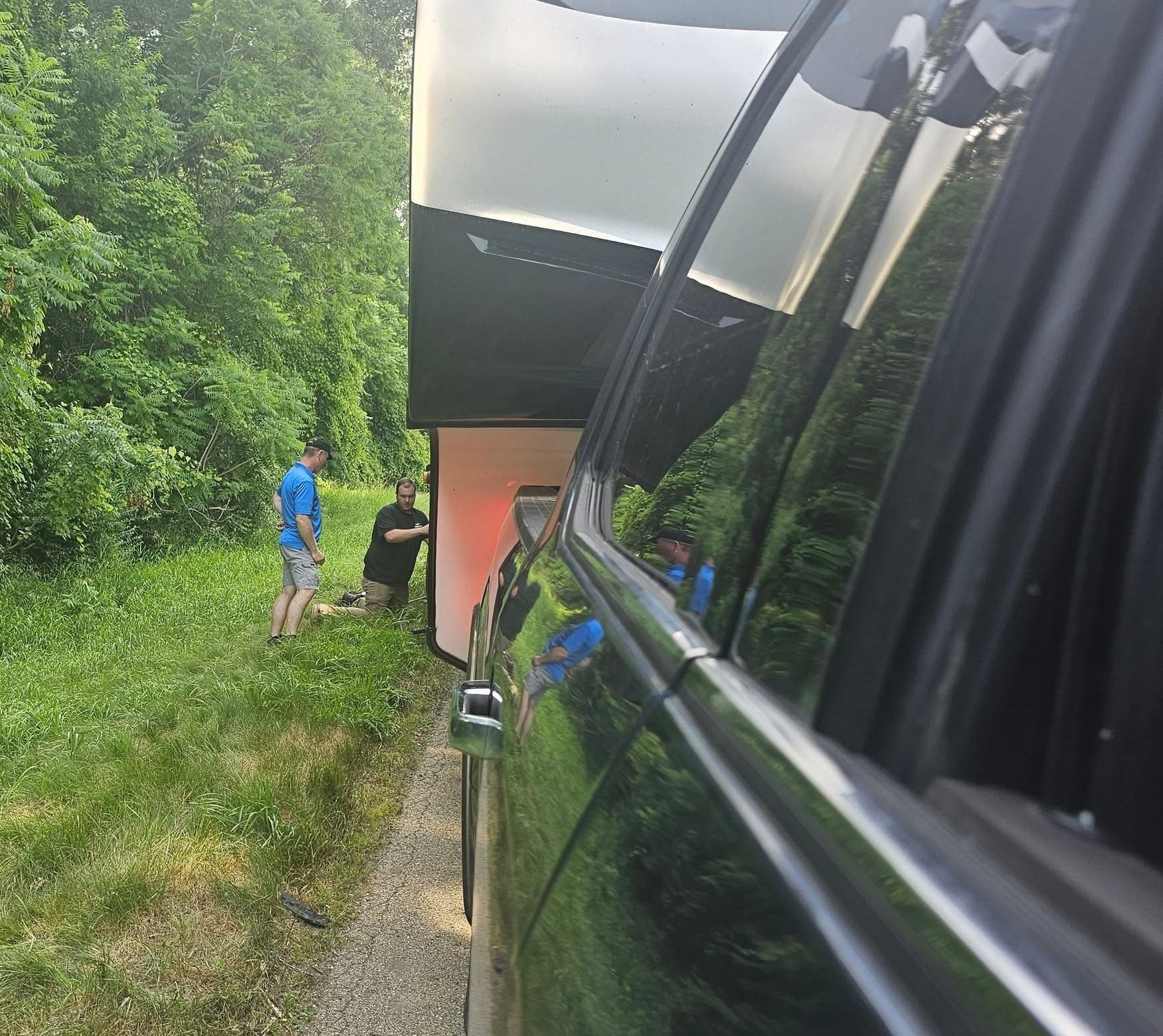 Two men near a trailer, alongside a car with reflective windows. Green trees and grass form the backdrop.