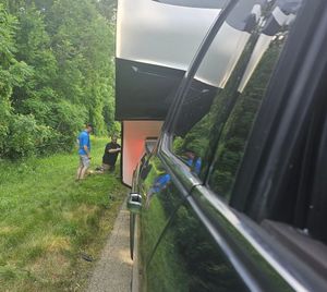 Two men near a trailer, alongside a car with reflective windows. Green trees and grass form the backdrop.