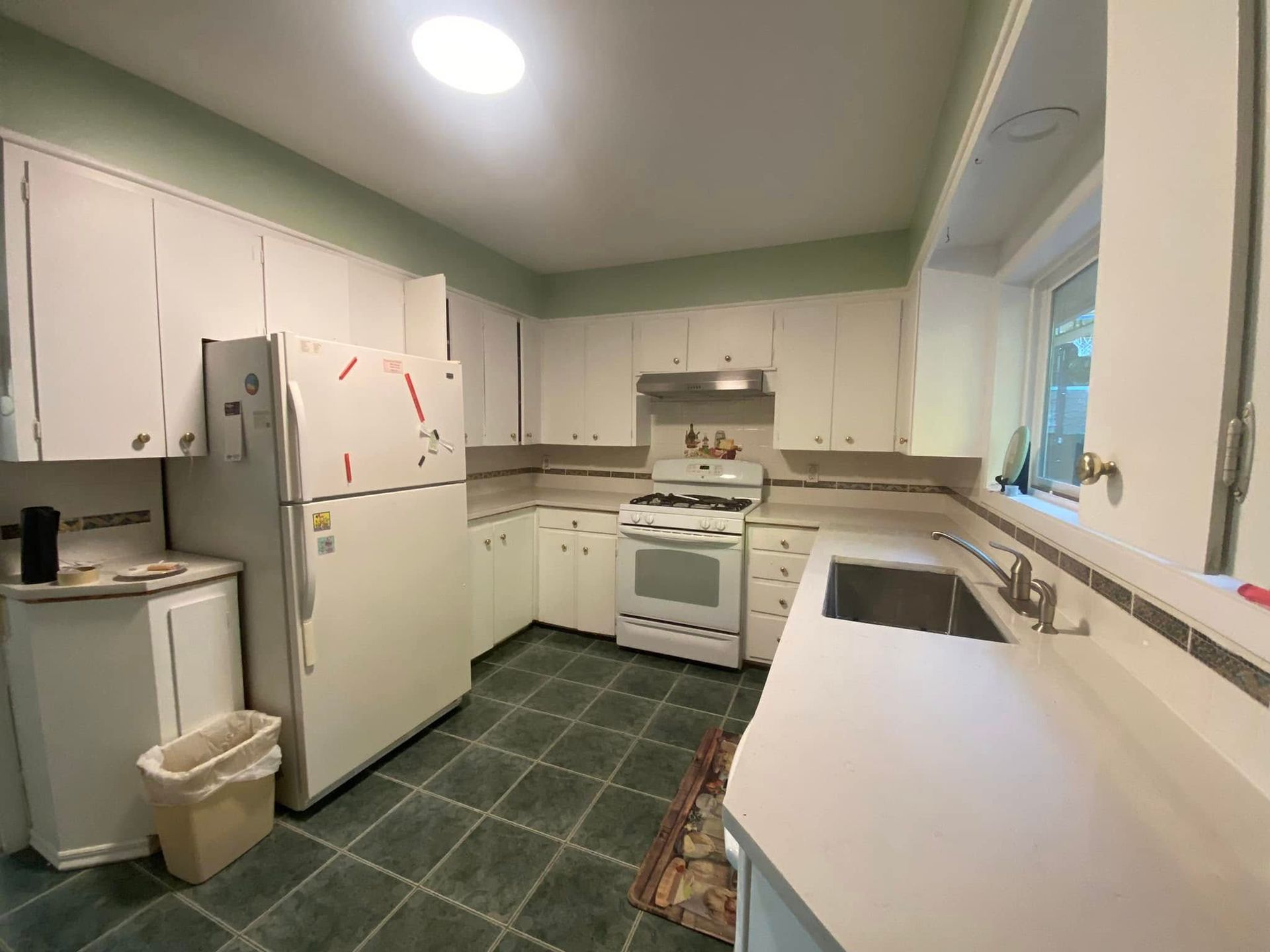 Small white kitchen with refrigerator, stove, cabinets, and a long counter by a window.