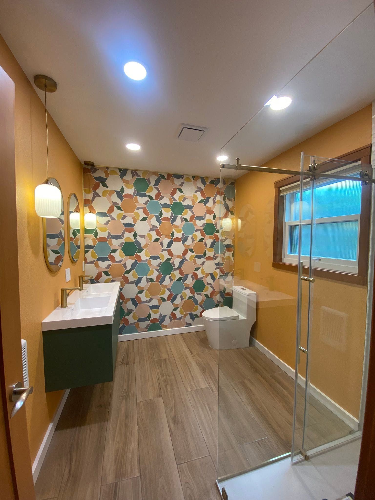 Modern bathroom with patterned accent wall, double sink vanity, toilet, and glass shower partition.
