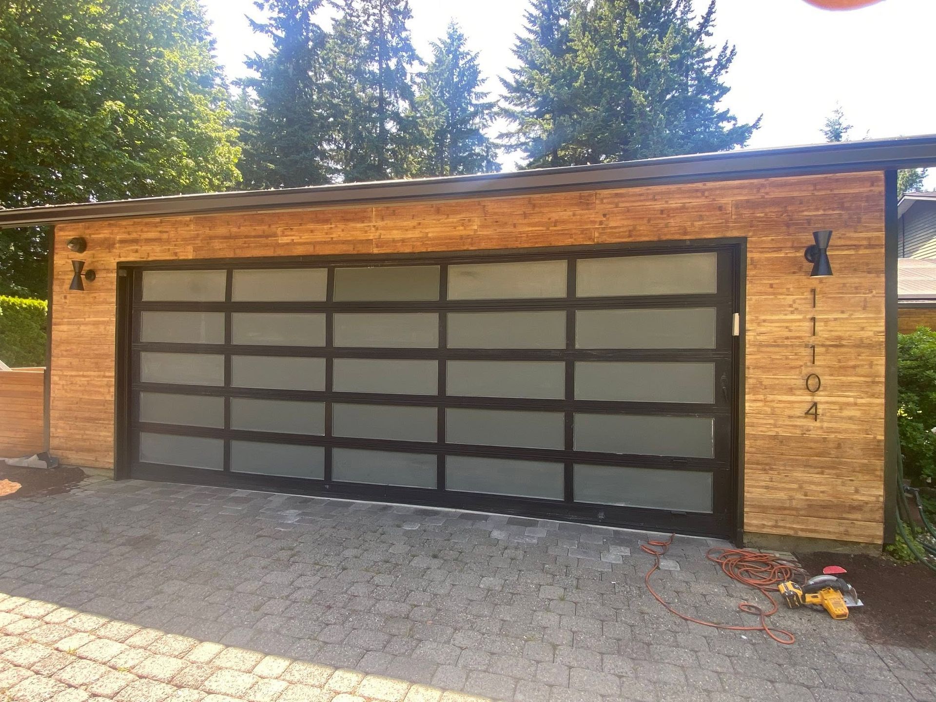 Single-story wooden garage with frosted glass doors and a paved driveway, framed by tall pine trees.