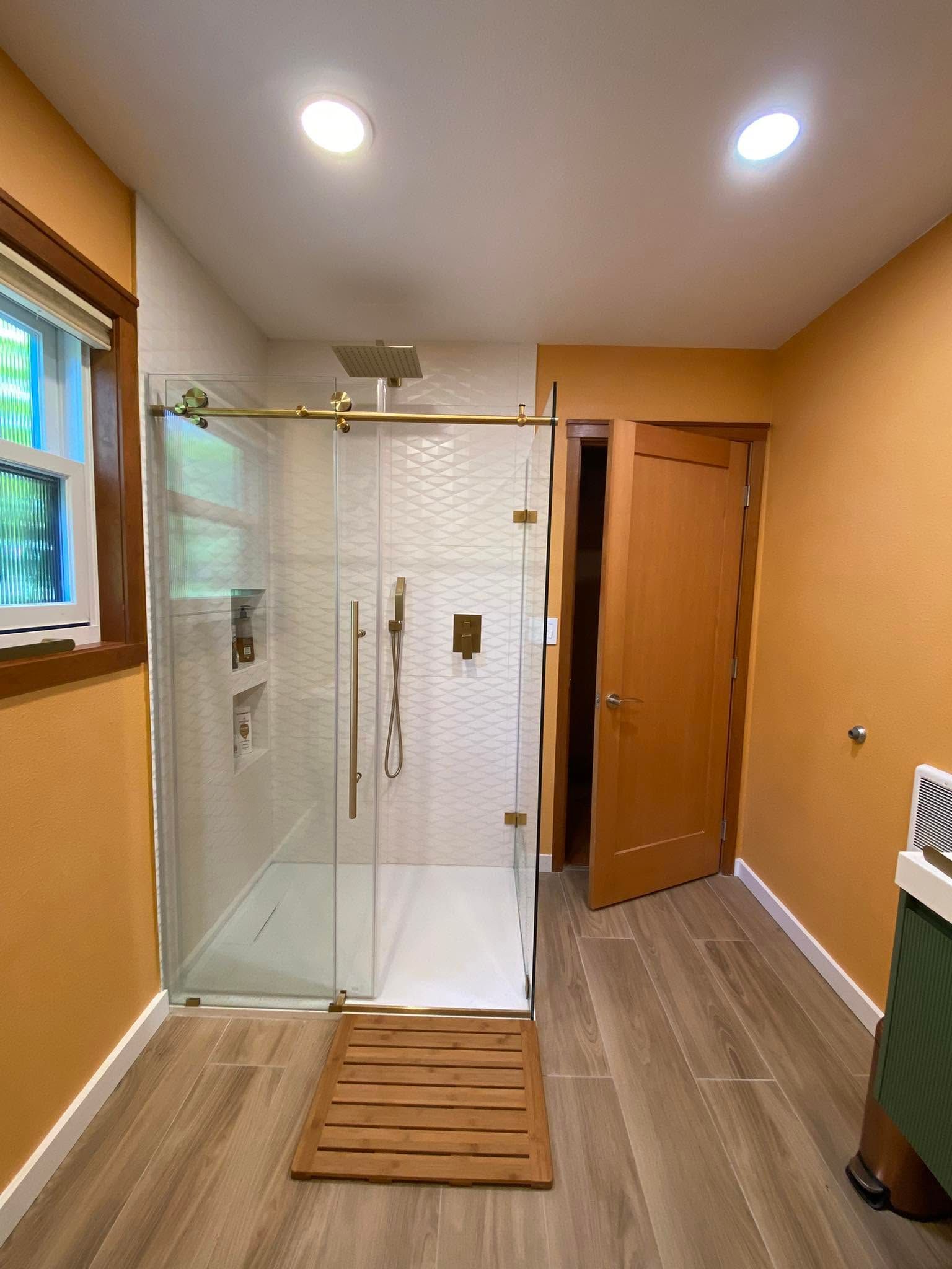 Bathroom with glass shower enclosure, tan walls, wood floor mat, and open door to a hallway.