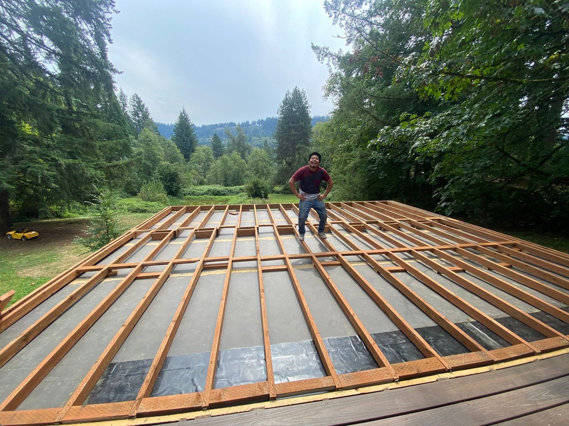 Person kneels on a wooden frame over a black tarp in a forest clearing, building a deck.