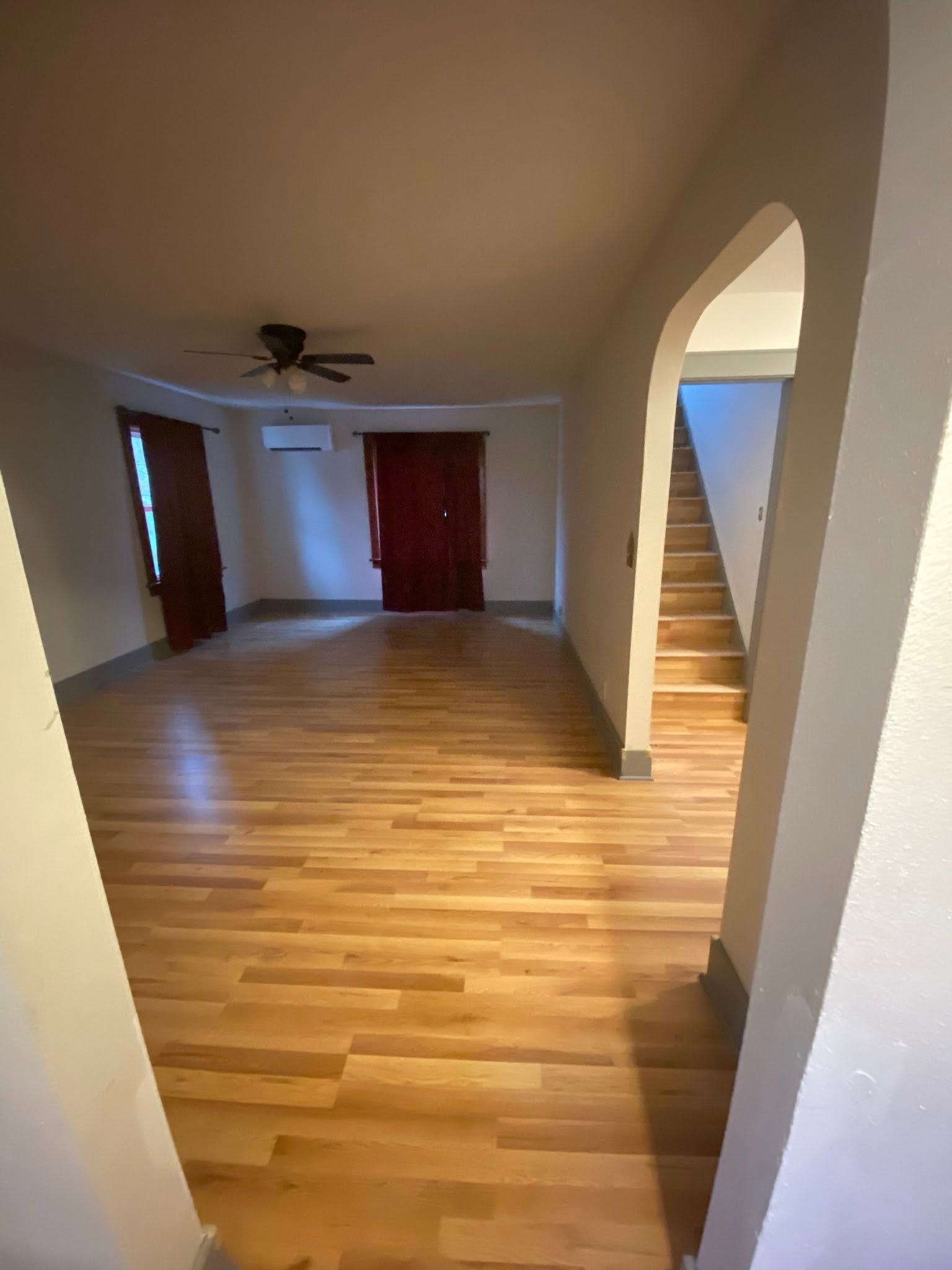 Empty living room with hardwood floors, ceiling fan, red curtains, and arched doorway to stairs.