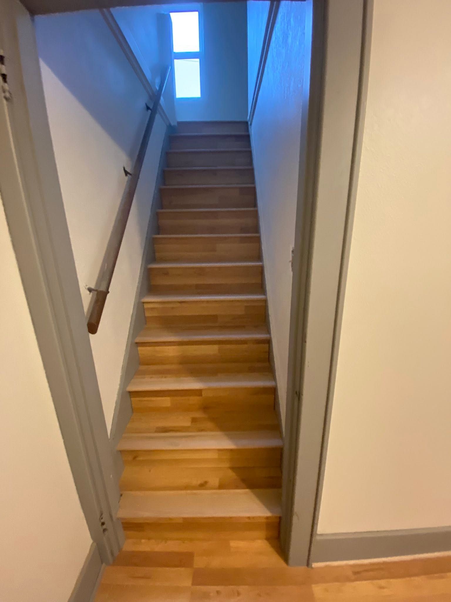 Narrow indoor staircase with wooden steps and handrails, lit by a bright window at the top.