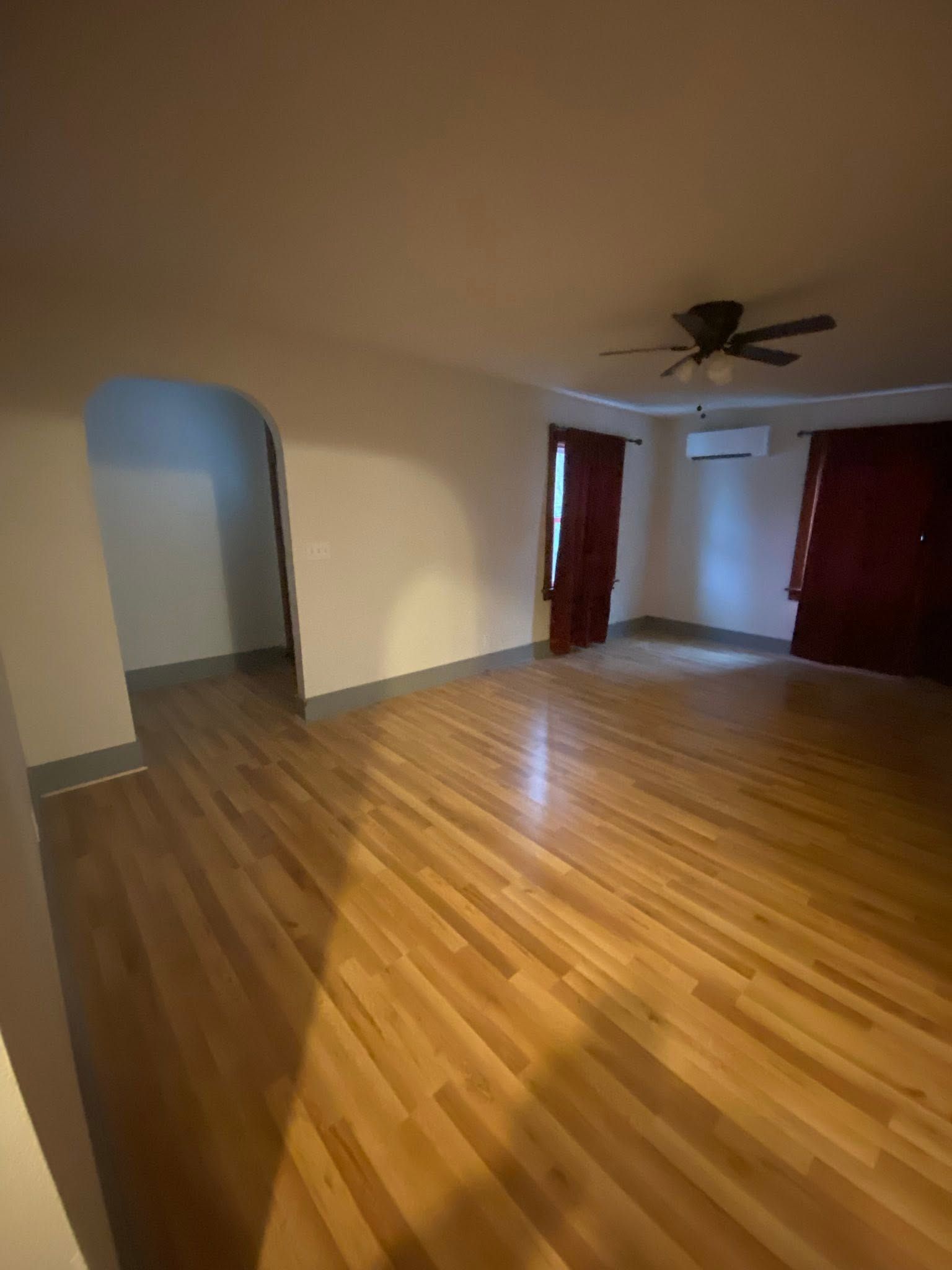 Empty living room with hardwood floors, beige walls, arched doorway, ceiling fan, and red curtains.