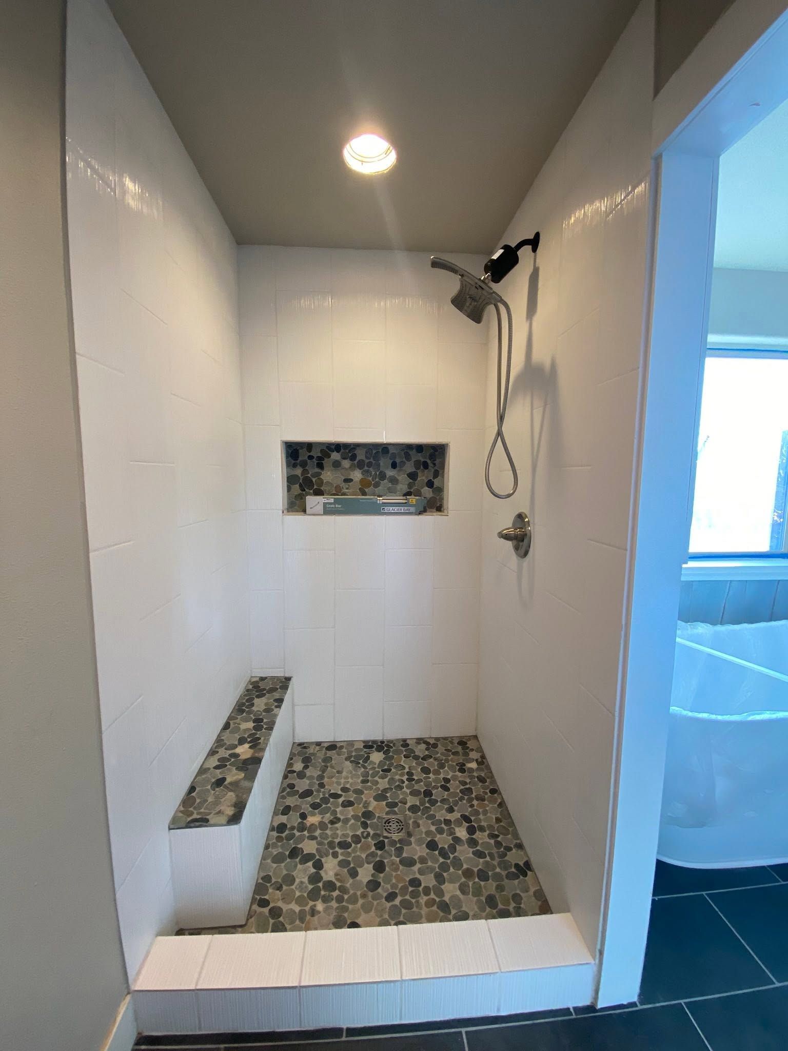 Small tiled shower stall with white walls, pebble floor, built-in bench, and chrome shower fixtures.