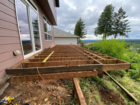 Deck framing under construction beside a house, overlooking a wooded hillside view.