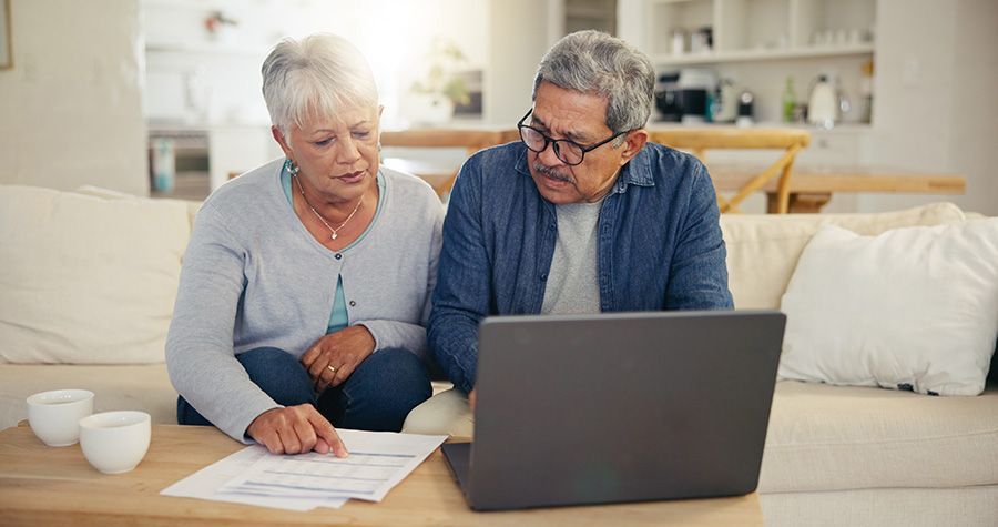Two people sitting on a couch at home, looking at a document and a laptop screen while discussing finances.