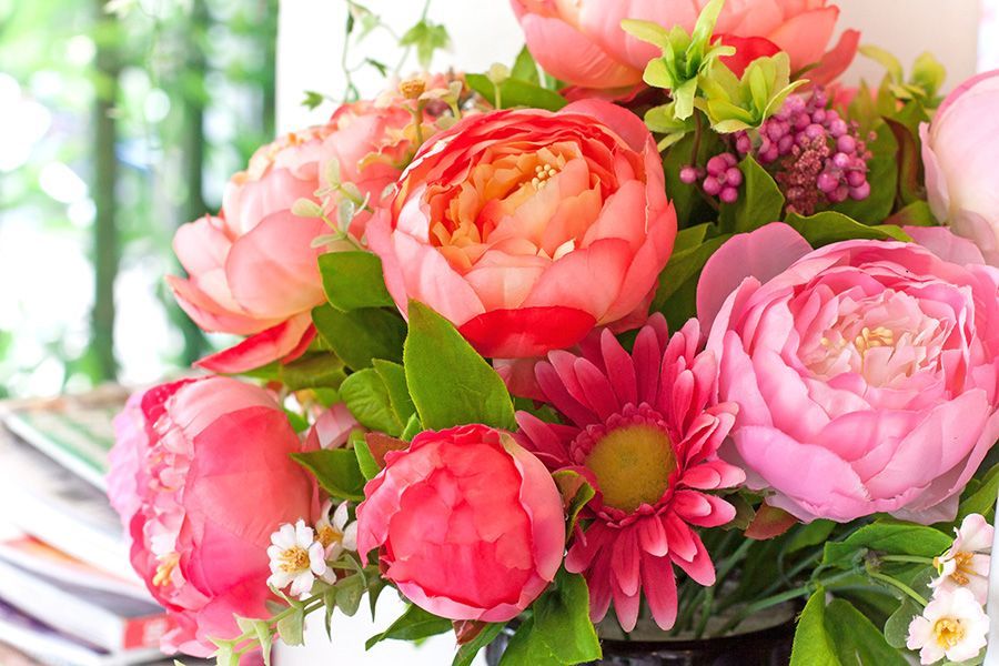 A lush bouquet of pink, coral, and red peonies with green leaves and small white flowers in a bright room.