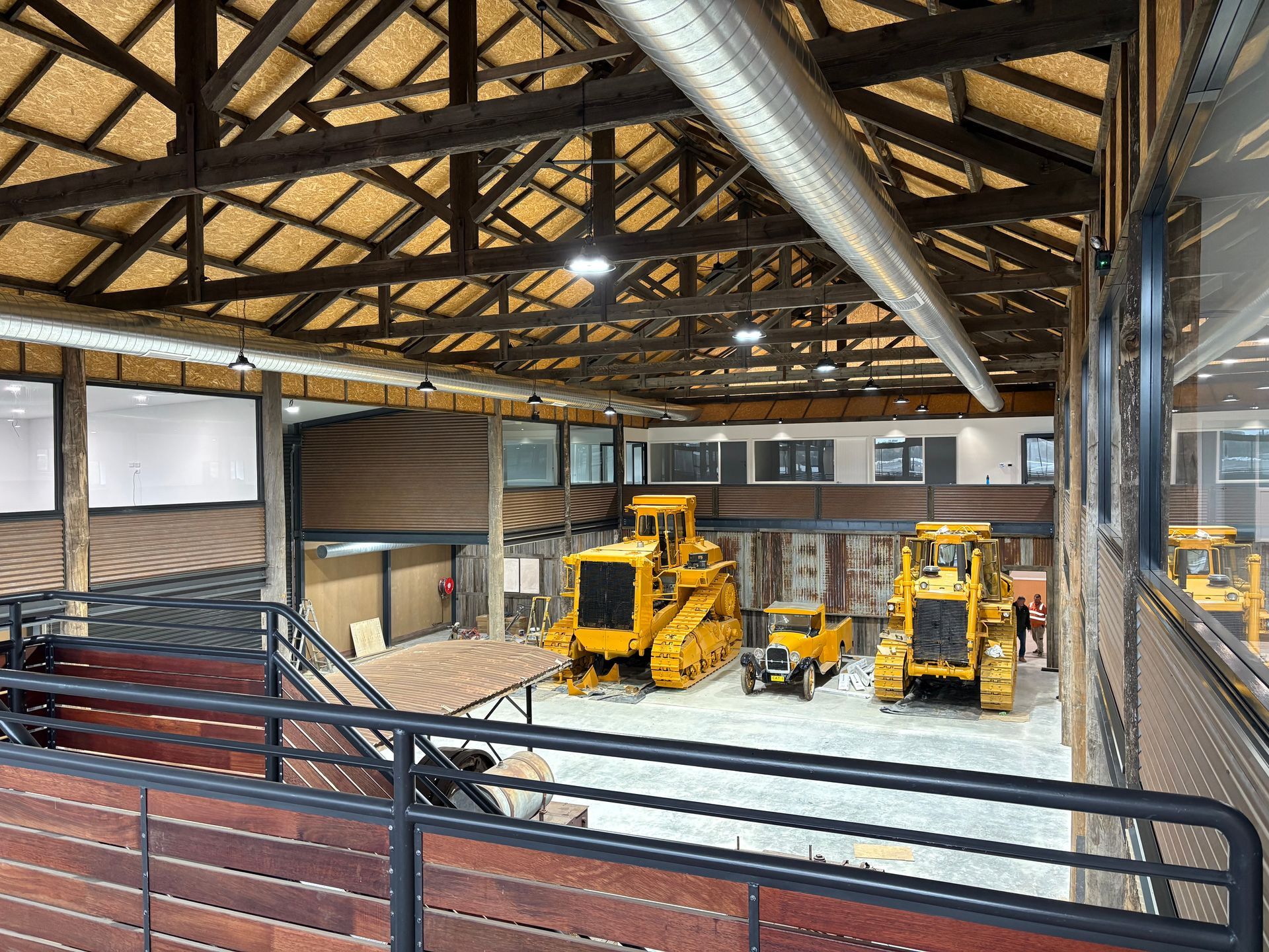Indoor space with several yellow construction vehicles on display. Brown wooden beams, dark railings, and concrete floor.