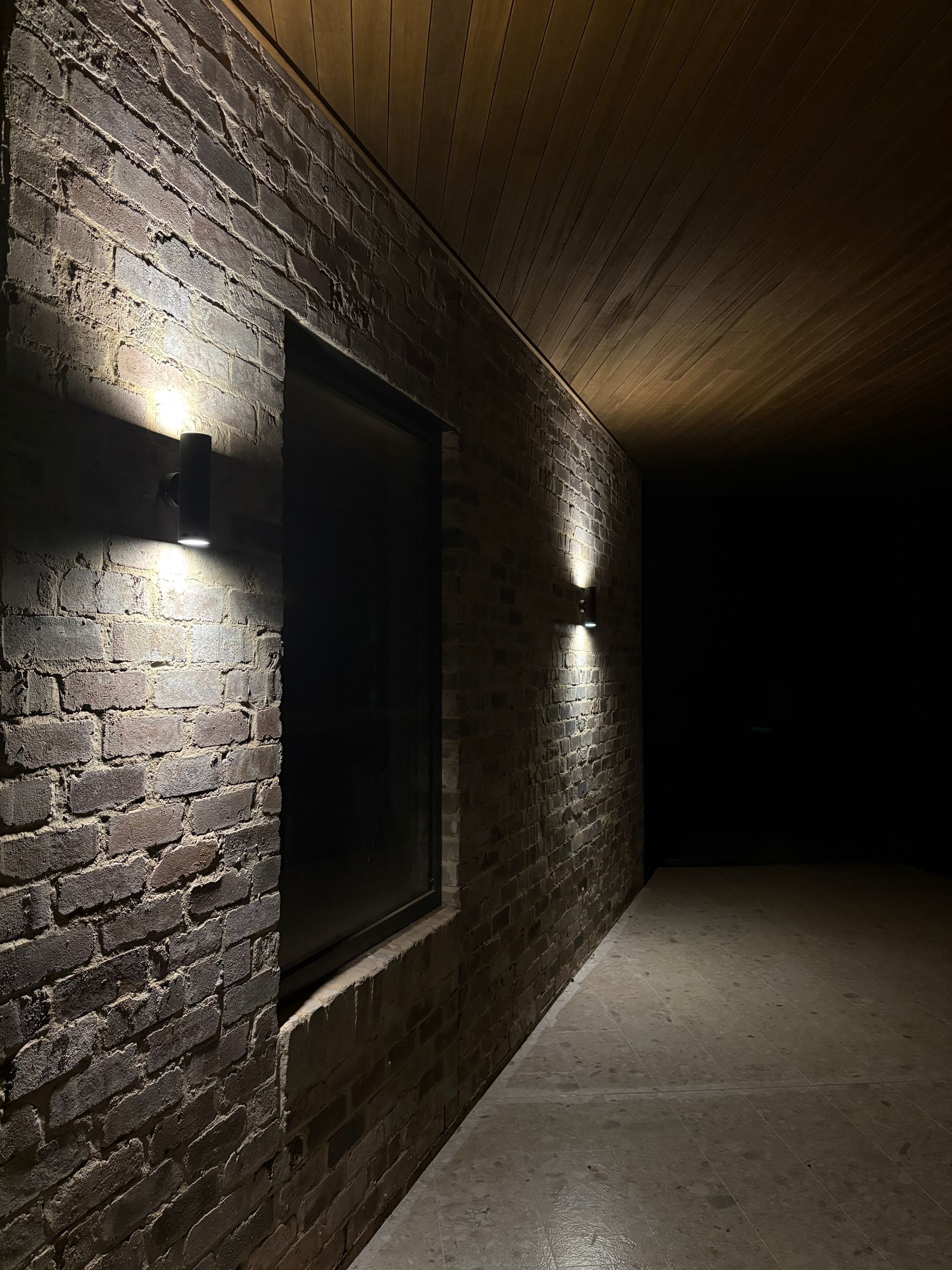 Brick-walled hallway illuminated by spotlights. Window on left side; dark space at end.