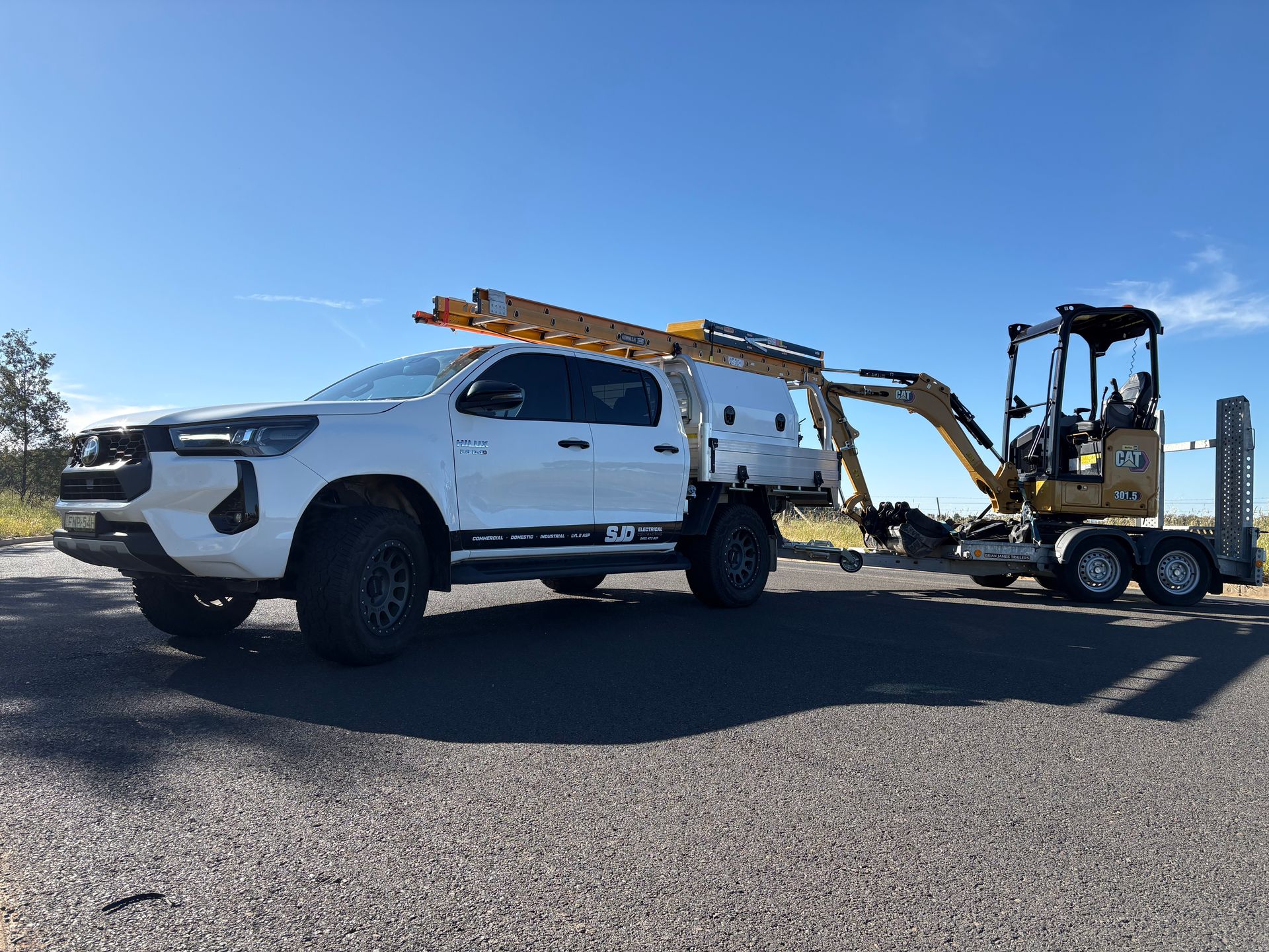 White truck hauling a trailer with a small excavator on a gravel road, under a blue sky— SJD Electrical Contracting in Orange, NSW