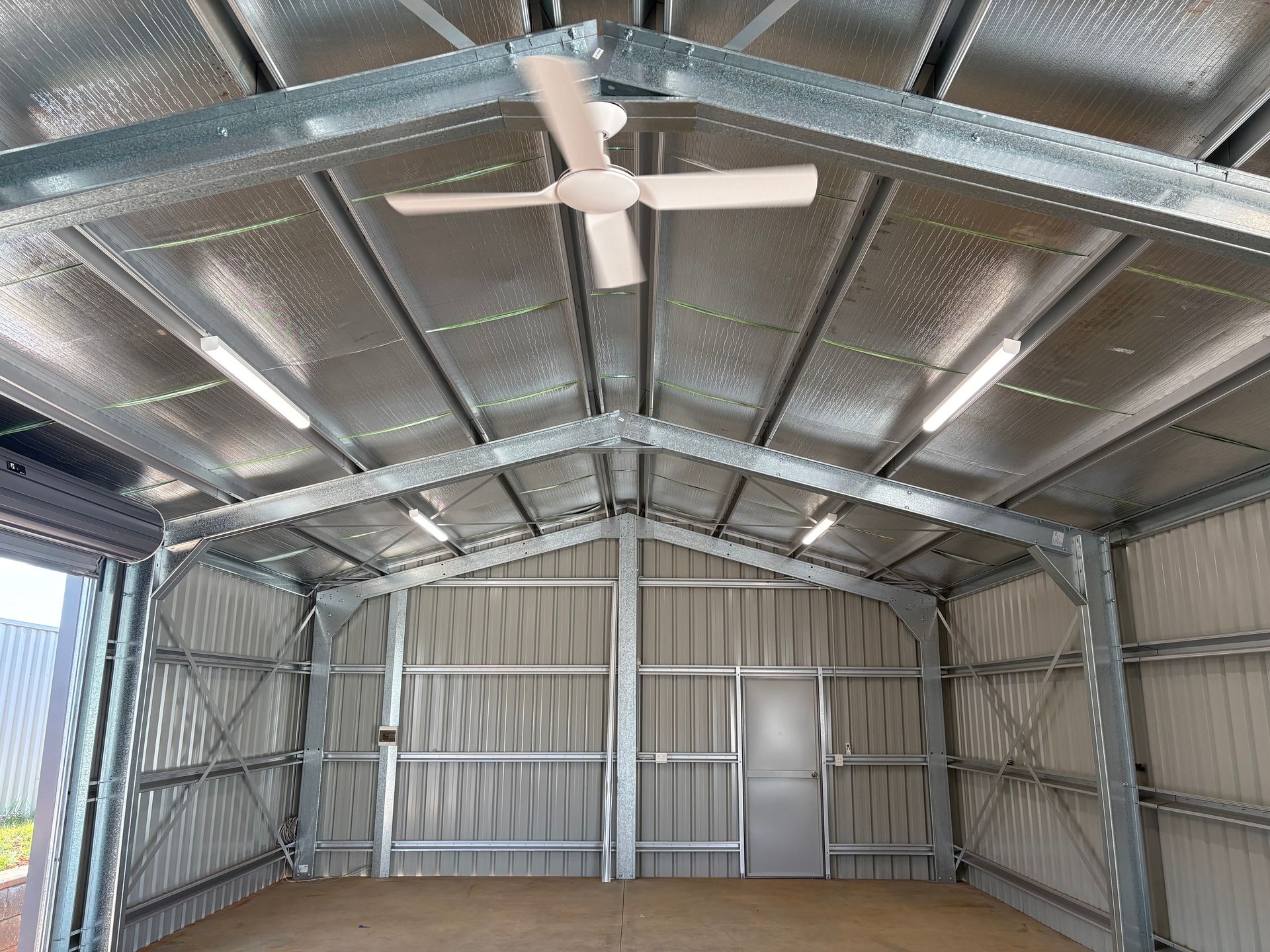 Interior of a metal shed with exposed beams, insulation, lights, ceiling fan, and a door.