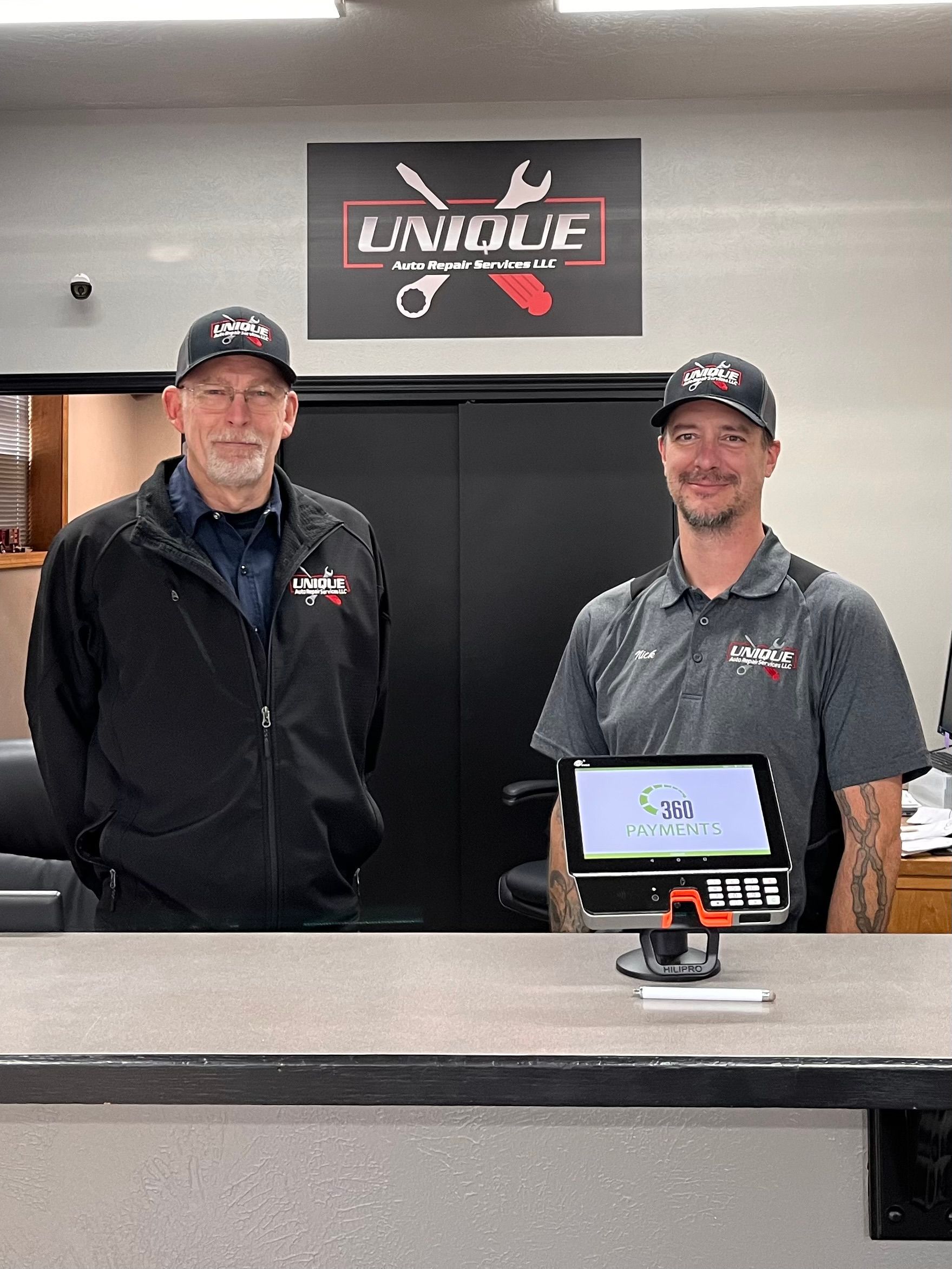 Two men behind a counter at Unique Auto. One is wearing a black jacket; the other has a gray shirt and tattoos, with a tablet in front of them.