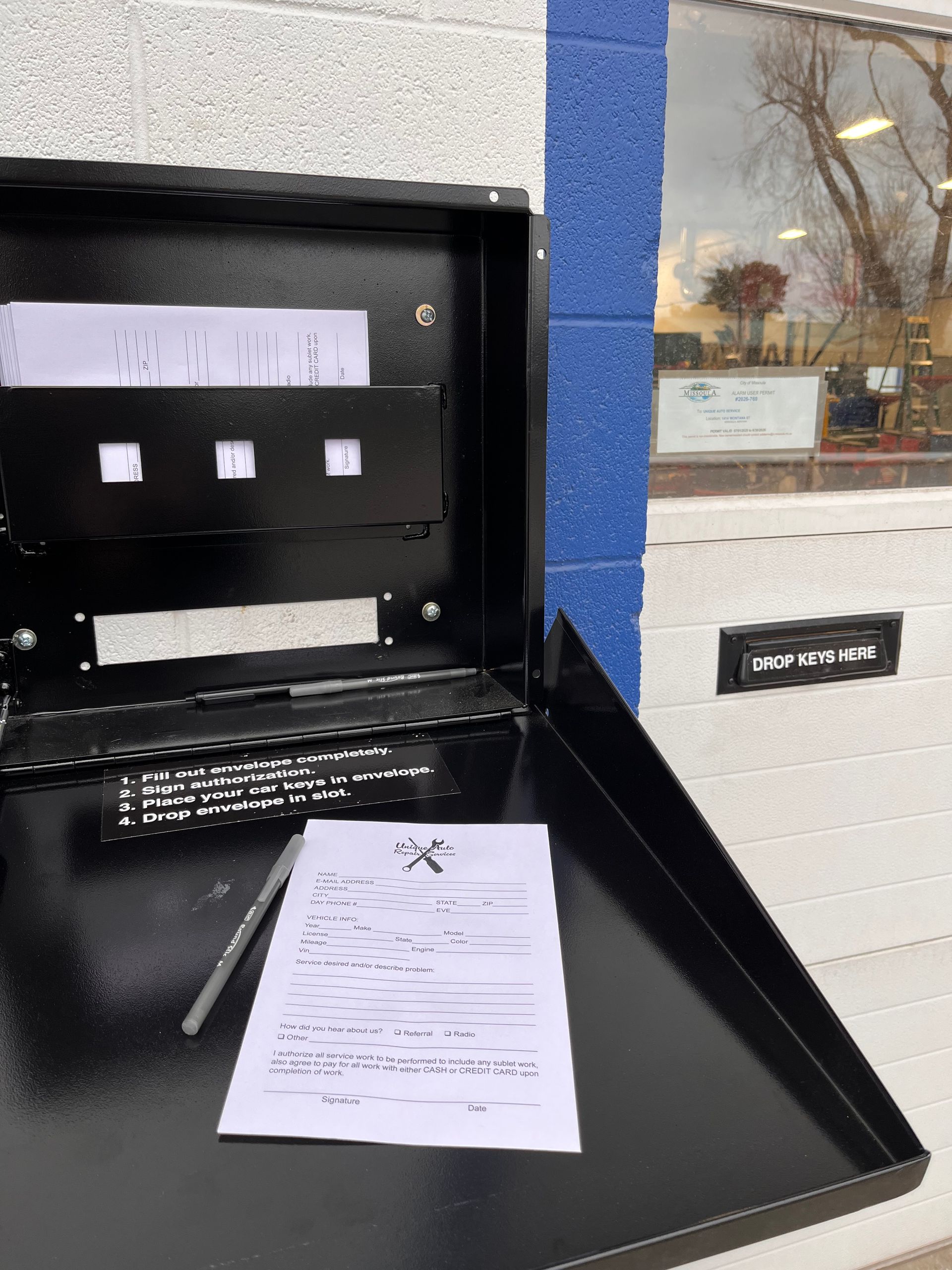 Black drop box with open door; a form and pen sit on the door, paperwork inside.