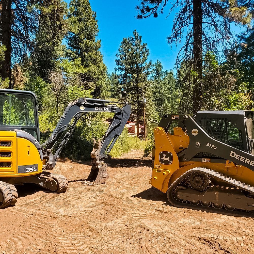 Yellow John Deere excavator and skid steer on a dirt clearing in a forest, digging on a sunny day.