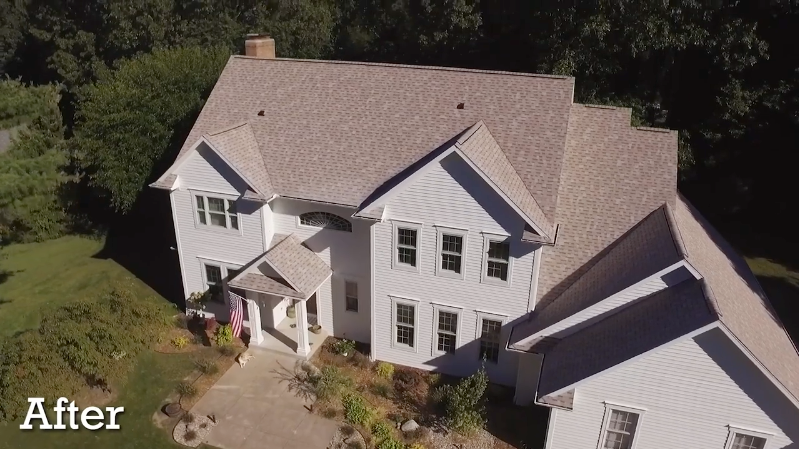 An aerial view of a two-story white house with a tan shingled roof, surrounded by trees and a paved front walkway.