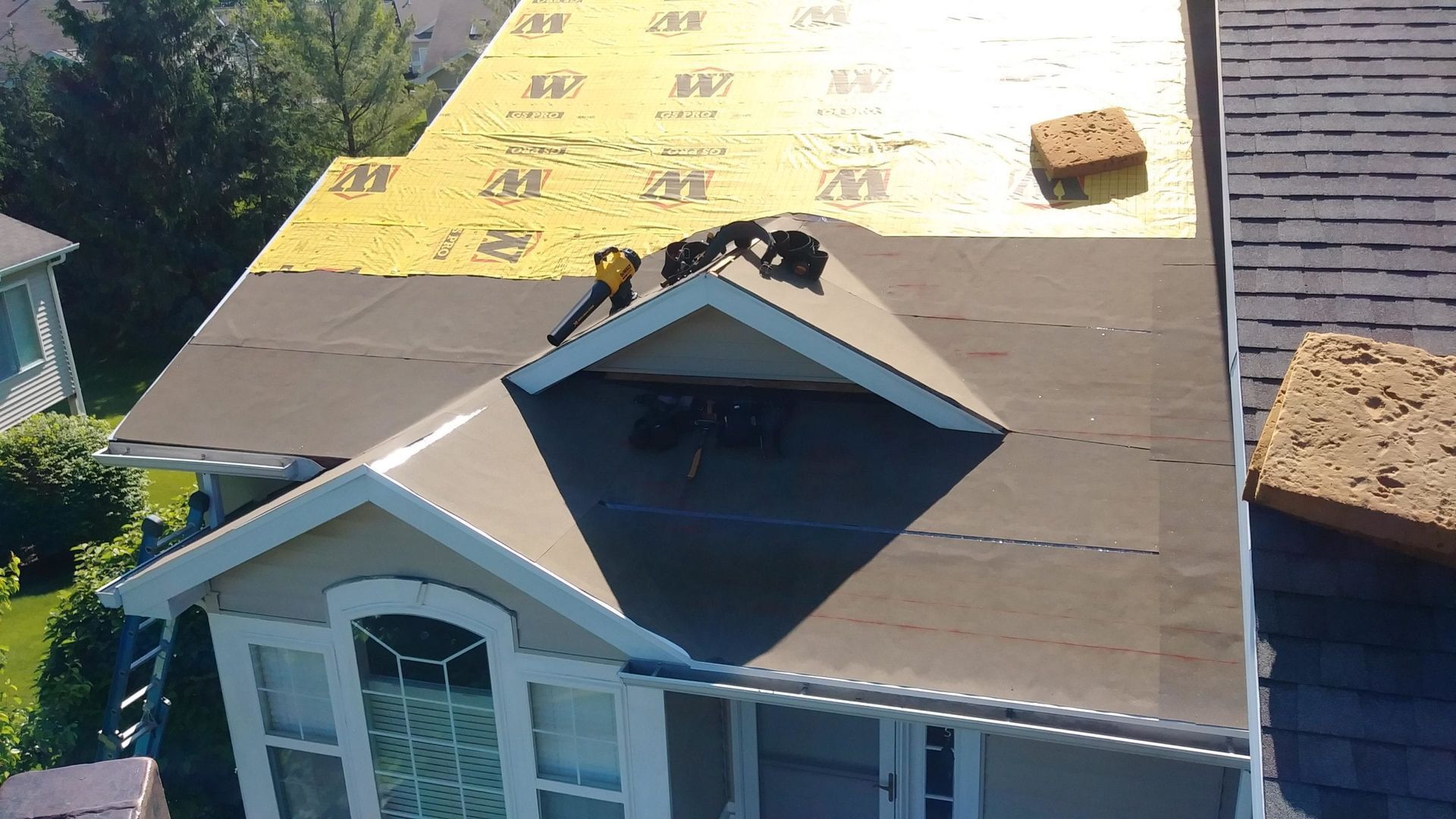 A worker stands on a partially shingled residential roof with sections covered in yellow underlayment.