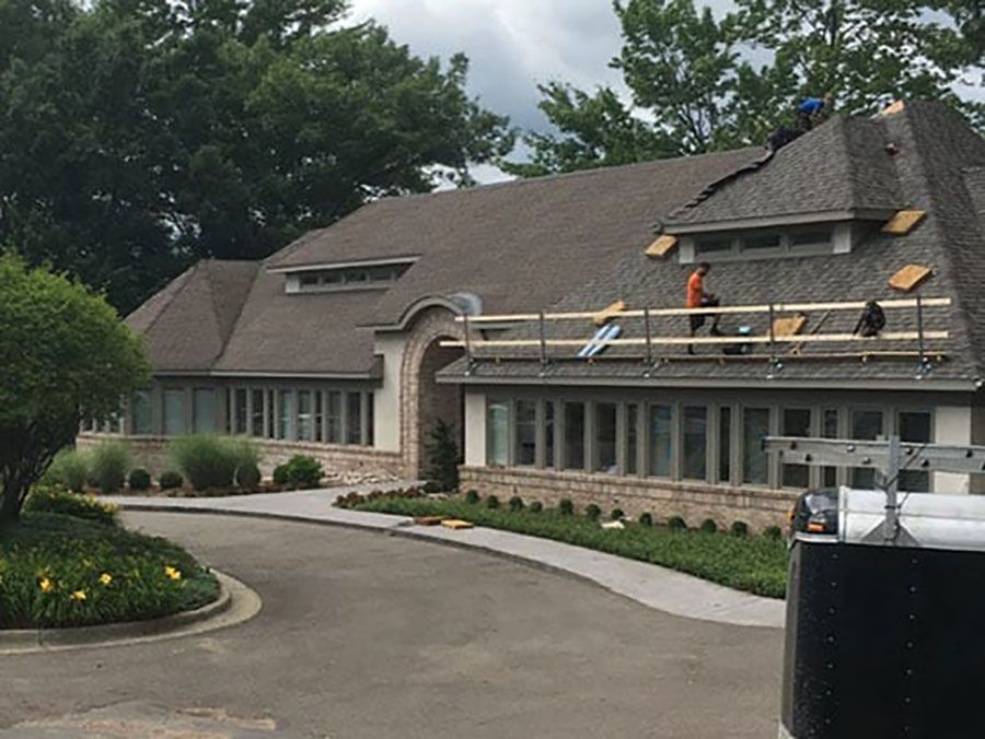 Workers on a safety scaffold repair the shingled roof of a suburban commercial building with a circular driveway.
