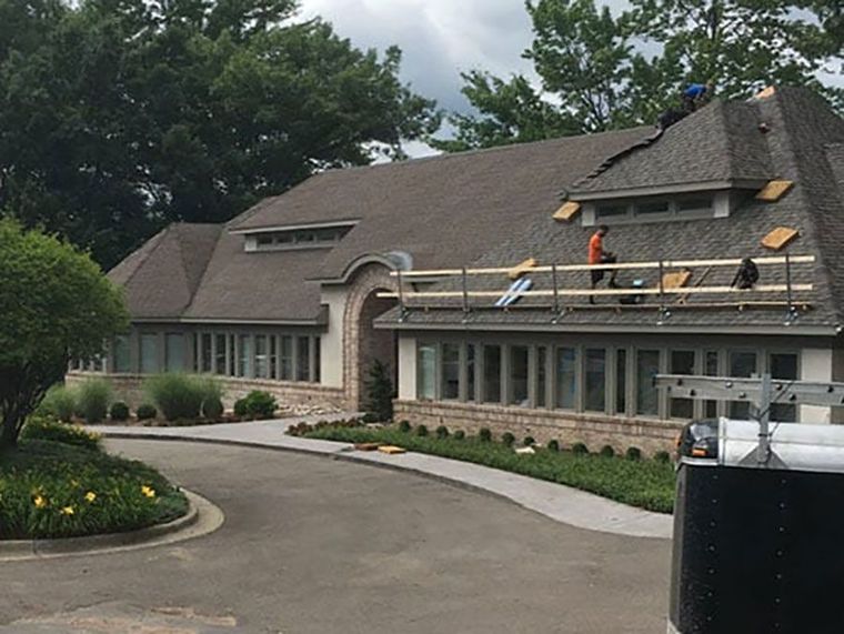 Workers on a safety scaffold repair the shingled roof of a suburban commercial building with a circular driveway.