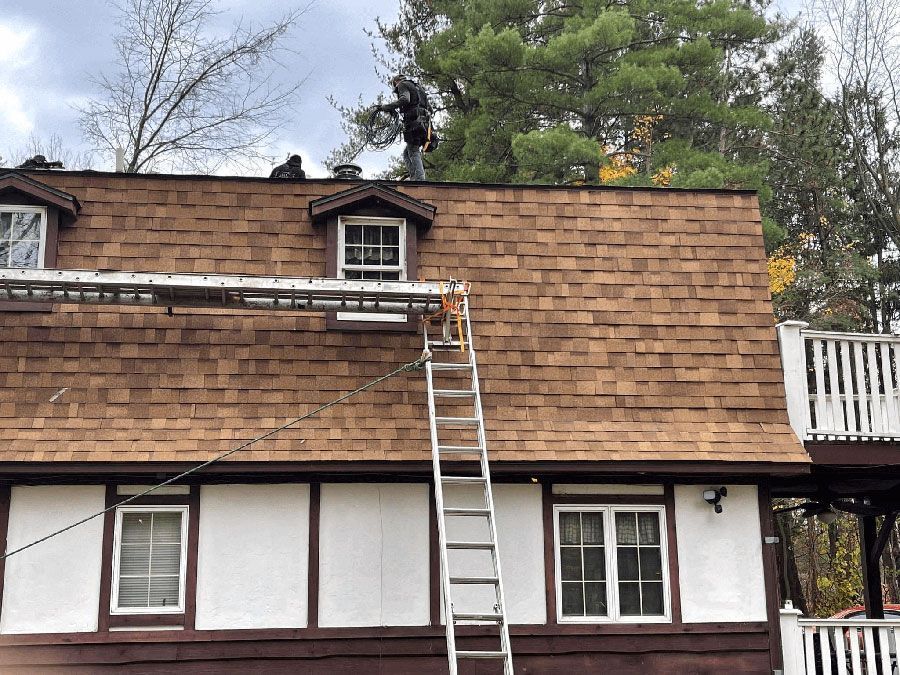 A person stands on the roof of a two-story home with brown shingles, preparing to work near a metal ladder.