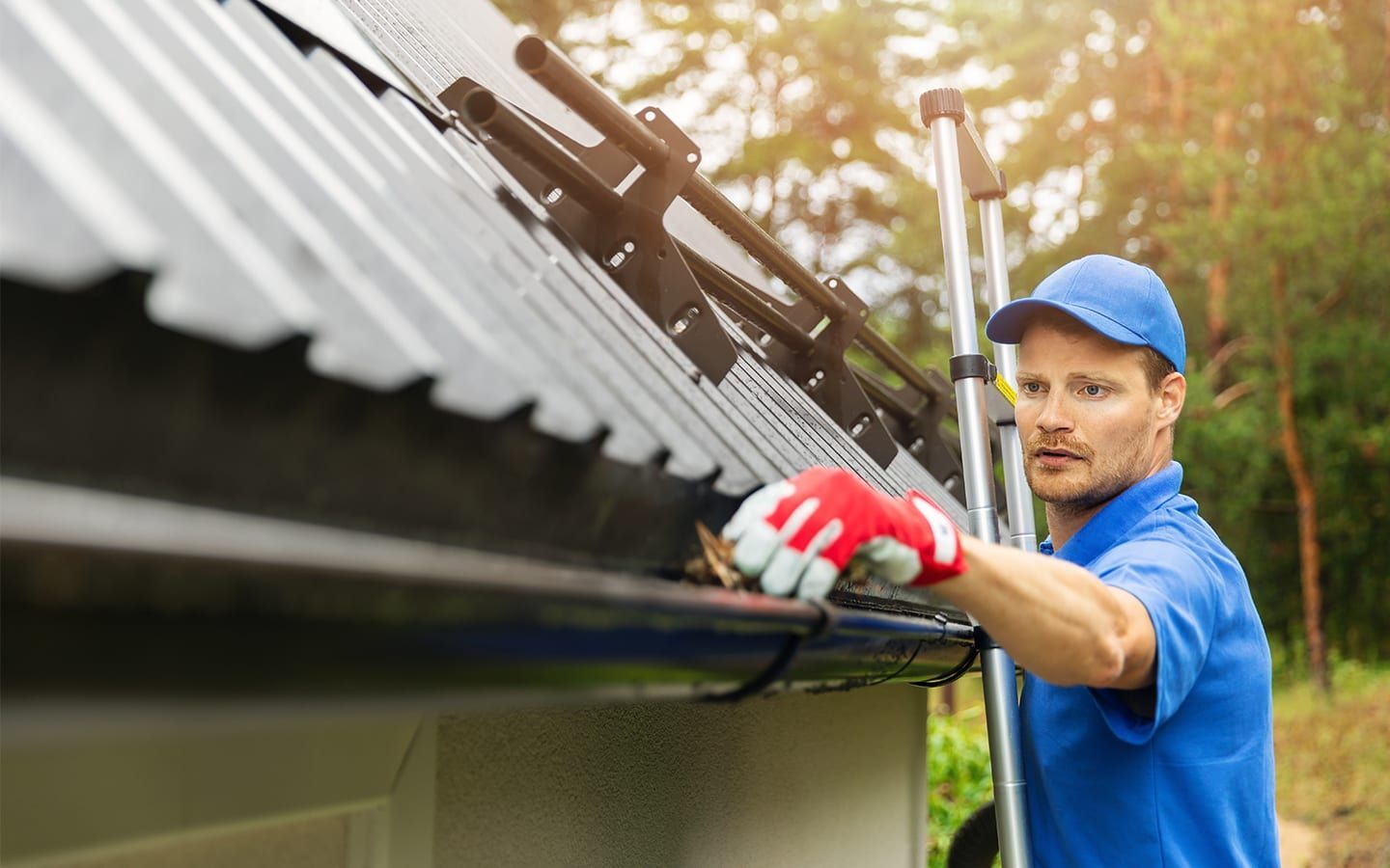A person in a blue uniform and cap cleans debris from a gutter while standing on a ladder against a metal roof.