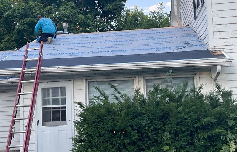 A person in a blue shirt works on a residential roof covered in blue underlayment, with a ladder leaning against the house.