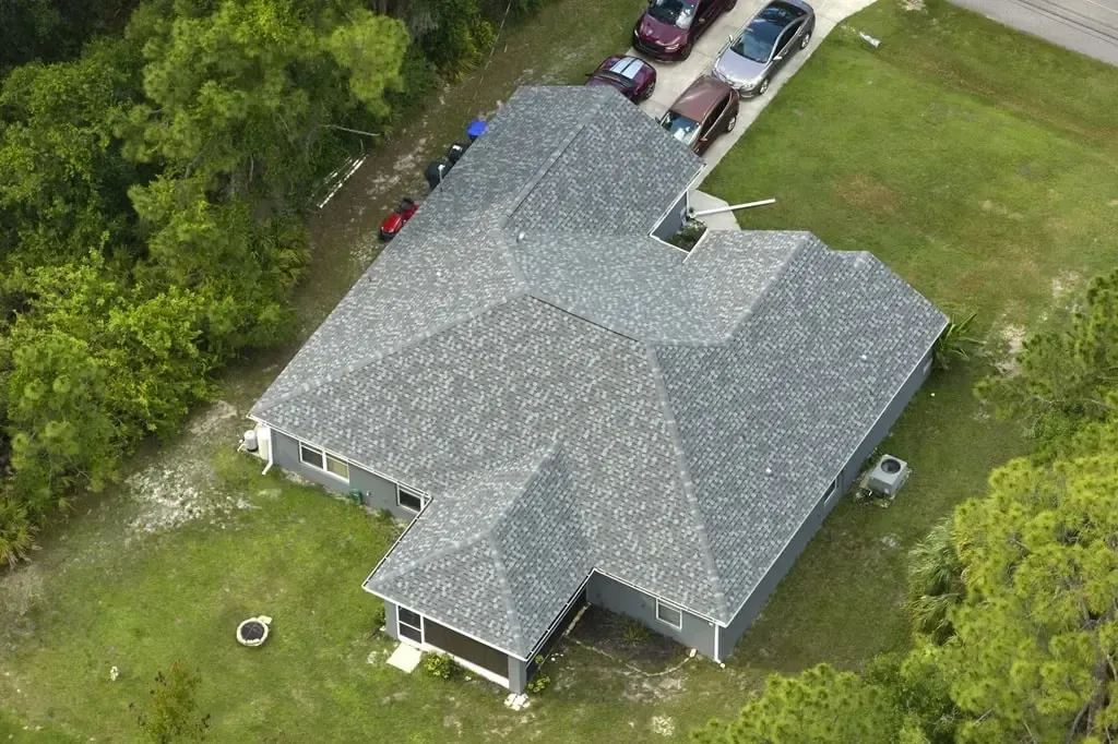 An aerial view of a suburban house with a gray shingle roof, surrounded by trees and a lawn with cars parked in the drive.