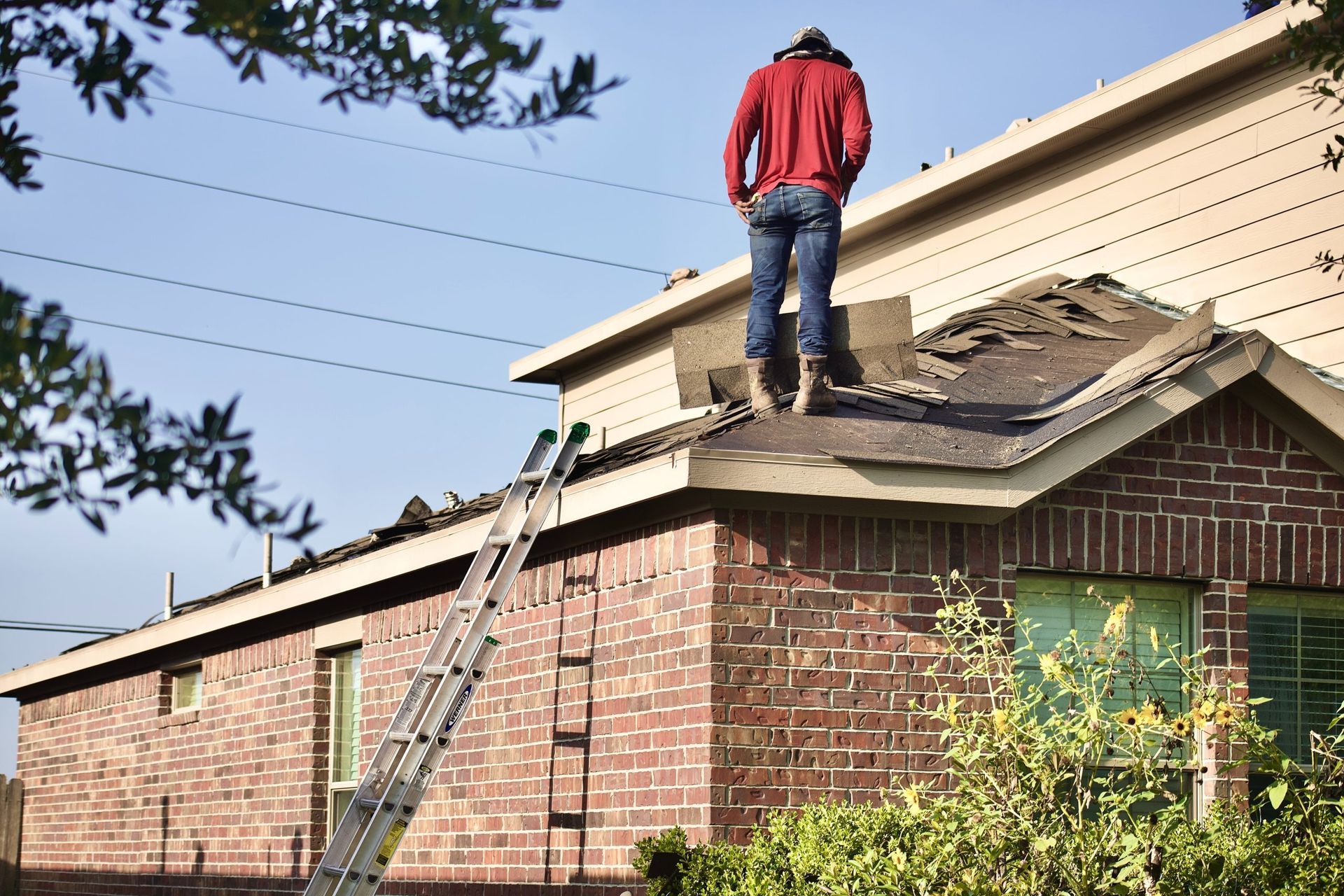A person in a red shirt and jeans stands on the roof of a brick house, performing repair work near a ladder.