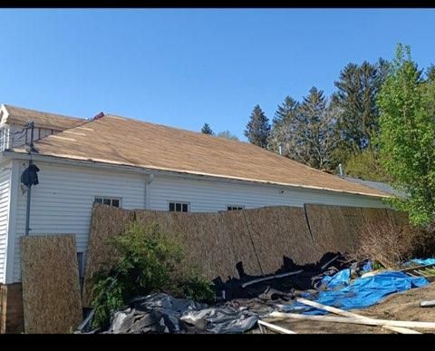 A side view of a white, single-story house with a newly sheathed roof and plywood boards covering the lower exterior wall.