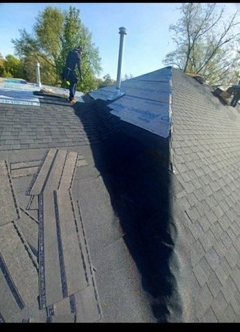 A worker installs gray shingles on a roof around a metal chimney pipe on a sunny day.