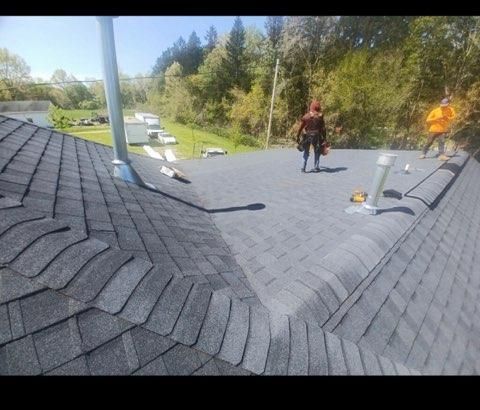 Two people in high-visibility gear work on a residential roof featuring gray shingles and multiple vent pipes.