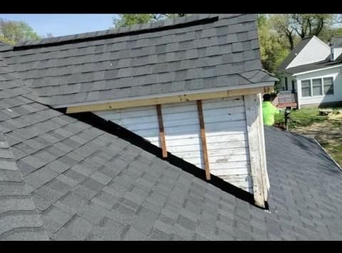 A person in a neon shirt works on a roof where new shingles meet an unfinished dormer with exposed wooden support beams.