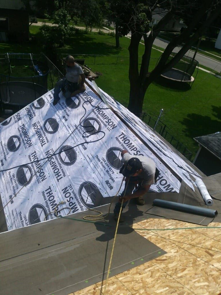 Two workers install synthetic underlayment on a plywood roof deck of a house under bright, sunny conditions.