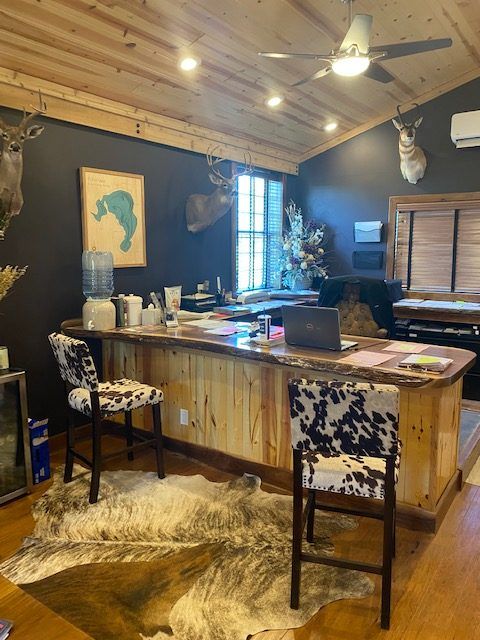 A rustic office with a live-edge wood desk, cow-print stools, a cowhide rug, and taxidermy mounts on dark blue walls.