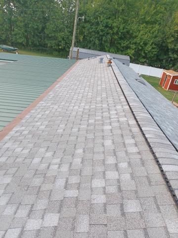A view looking down the roof ridge, showing grey asphalt shingles, a metal roof section, and a distant backyard.