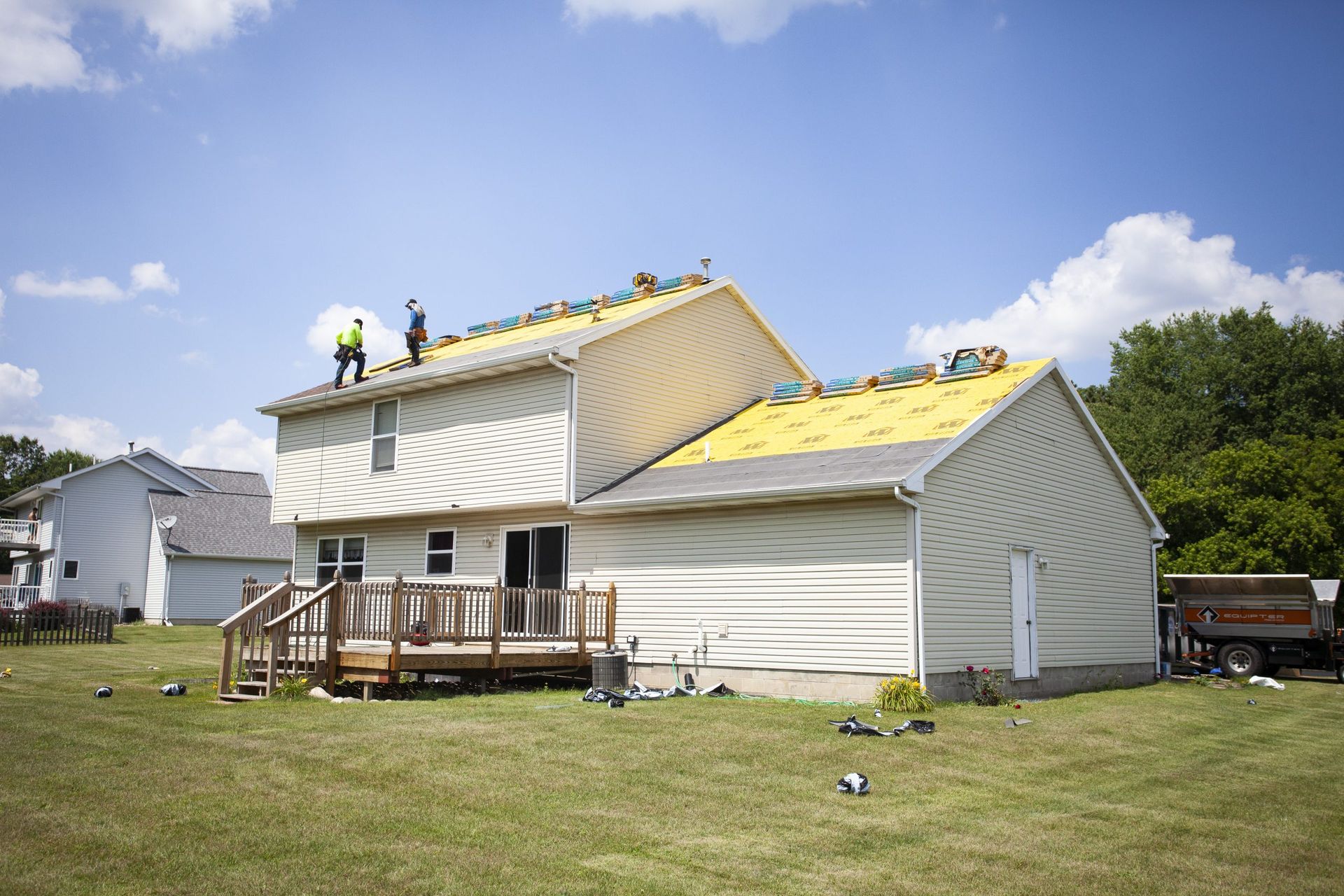 Workers replace the roof of a two-story beige house on a sunny day.