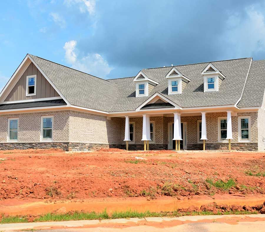 A newly constructed one-story brick house with a gray shingle roof, white columns, and a large red-dirt front yard.
