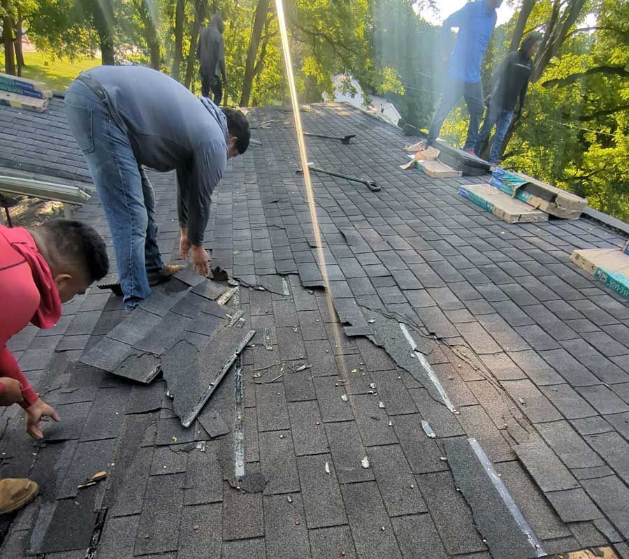 Three people work on a sloped roof, removing and replacing dark gray asphalt shingles in a sunny outdoor setting.