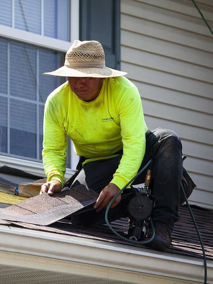 A person wearing a wide-brimmed straw hat and neon yellow long-sleeved shirt installing asphalt shingles on a roof.