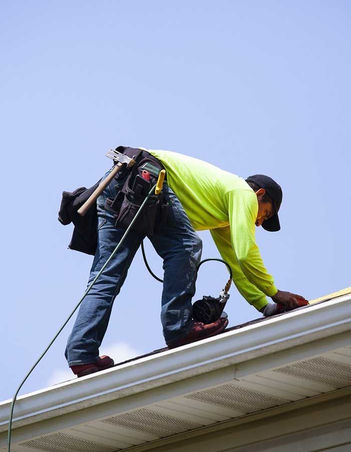 A roofer in a neon yellow long-sleeve shirt and tool belt works on a roof against a clear blue sky.