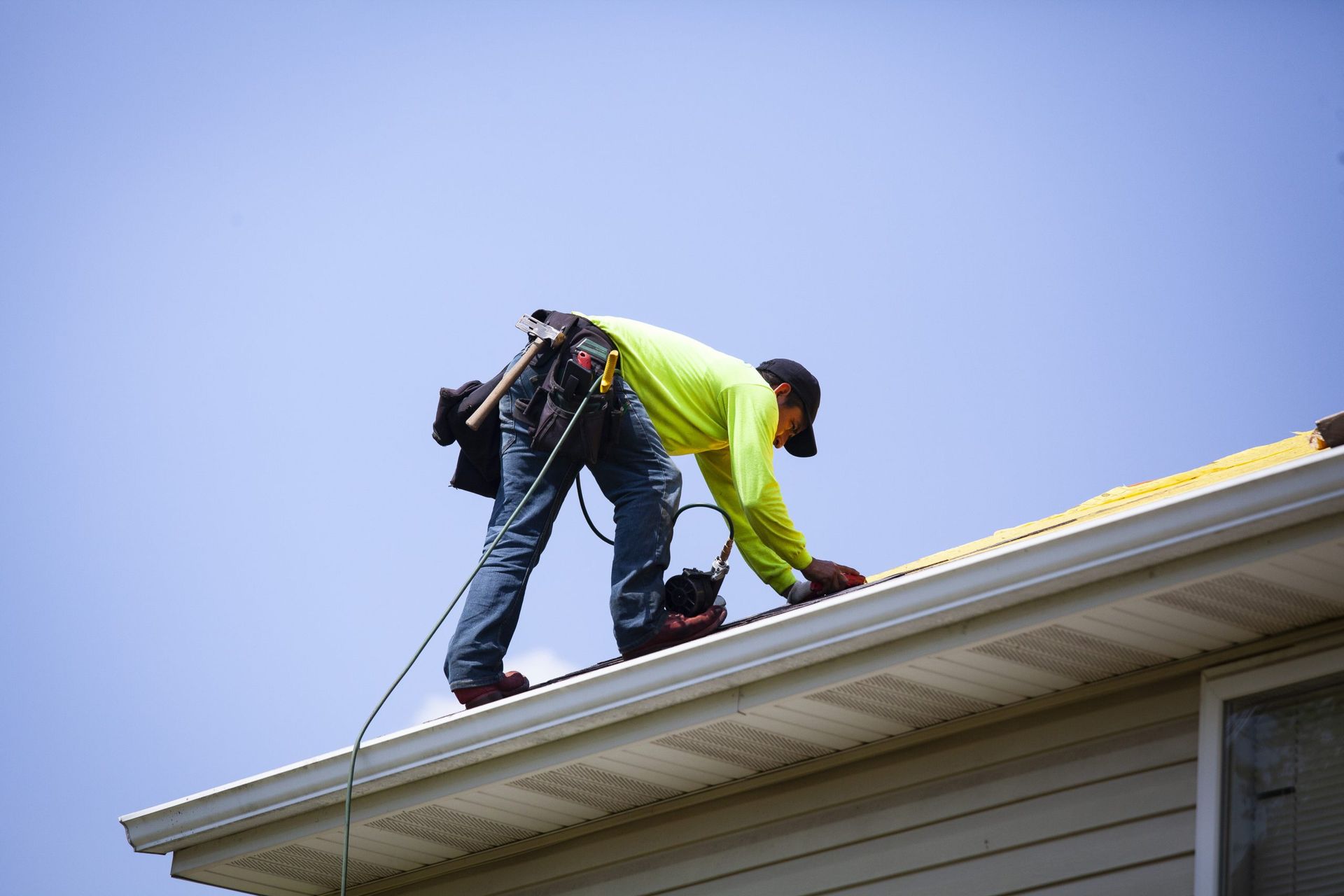 A roofer in a bright yellow long-sleeved shirt and tool belt working on the edge of a house roof against a blue sky.