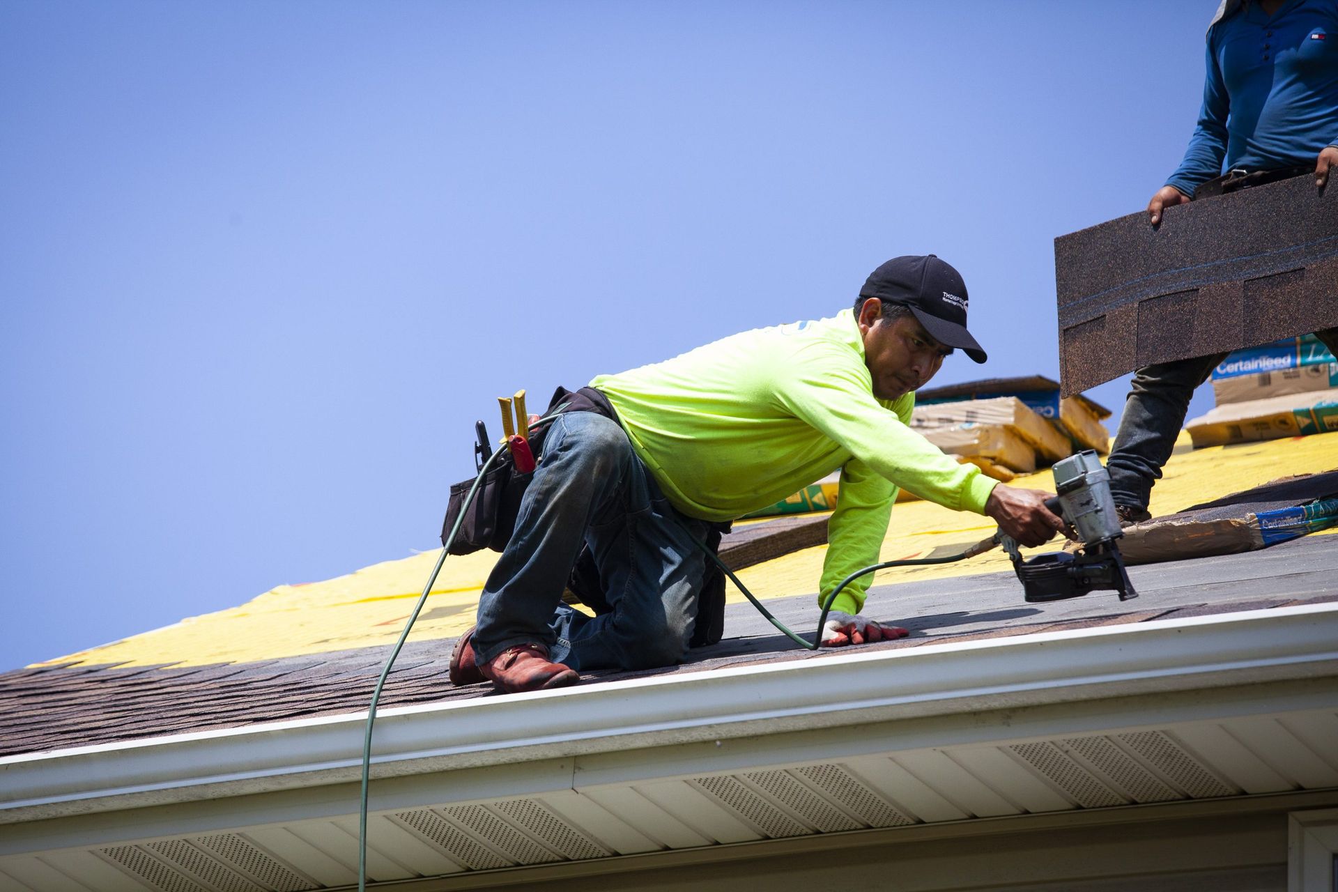 A roofer in a neon yellow shirt kneels on a residential roof, using a nail gun to install dark asphalt shingles.